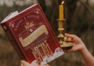 Na zdjęciu dłoń trzymająca książkę w czerwono-złotej okładce pod tytułem "Ostatni absolwent". Druga dłoń trzyma świecznik z zapaloną świecą. W tle naturalny krajobraz - drzewa, i fragment nieba.